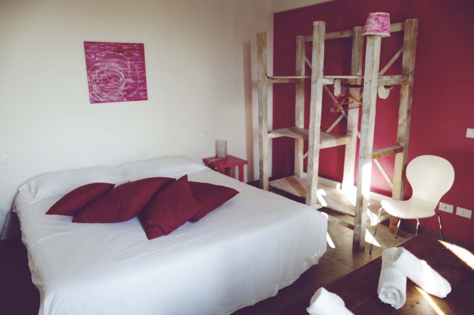 La Suite Lampone: letto con cuscini color lampone, parete fucsia, armadio aperto in legno di recupero e seduta di design bianca